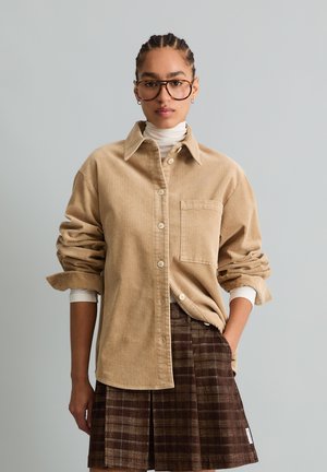 Donna con capelli intrecciati e grandi occhiali indossa una camicia di velluto a coste beige sopra un dolcevita bianco e una gonna scozzese marrone, in piedi davanti a uno sfondo semplice.