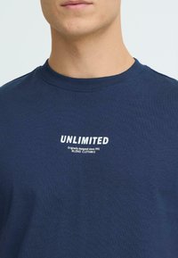 Laivastonsininen puuvillainen t-paita, jossa on pyöreä kaula-aukko ja valkoinen painettu teksti: "UNLIMITED" ja "Originally designed since 1993" rinnassa.