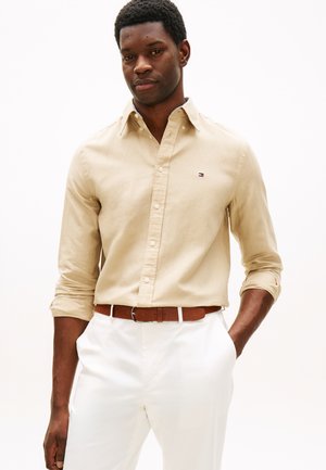 Mand iført beige skjorte med knapper og opkørte ærmer, brun bælte og hvide bukser, stående med den ene hånd i lommen mod en hvid baggrund.