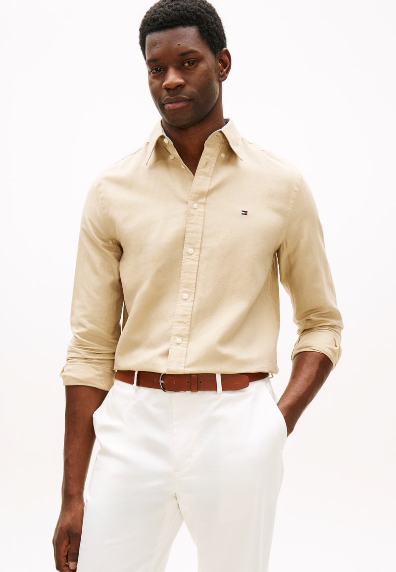 Άνδρας που φοράει μπεζ πουκάμισο με κουμπιά και γυρισμένα μανίκια, καφέ ζώνη και άσπρο παντελόνι, στέκεται με το ένα χέρι στην τσέπη μπροστά σε λευκό φόντο.