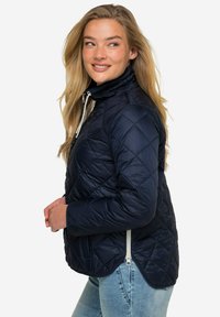 Marinblå kviltad jacka med hög krage, zippered framtill och sidofickor. Har en mjuk textur och en lätt rundad fåll.