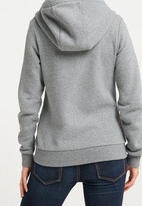Felpa grigia con cappuccio, dotata di polsini e orlo a coste, con una texture morbida e una tasca a marsupio, abbinata a jeans blu scuro.