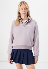 Blonde woman wearing a light purple quarter-zip fleece over a collared shirt and a navy blue pleated skirt against a white background.