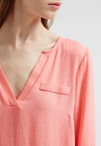 Blusa corallo realizzata in tessuto leggero; presenta un collo a V, maniche lunghe e una piccola tasca applicata sul petto. Texture liscia.