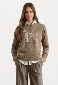 Beige strikkegenser med brodert hundedesign. Ribbede kanter og mansjetter, lagd over en hvit skjorte med krage.