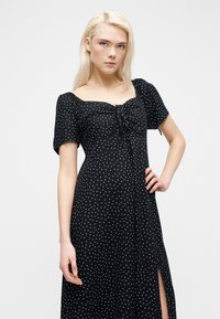 Νεαρή γυναίκα με ξανθά μαλλιά, φορώντας μαύρο φόρεμα με λευκές πουά, κοντά μανίκια και σχίσιμο μπροστά, όρθια με το χέρι στη μέση.