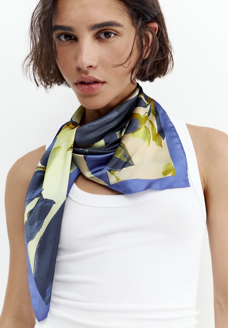 Femme aux cheveux courts et foncés portant un débardeur blanc et un foulard en soie coloré avec des motifs floraux bleus, jaunes et verts autour du cou.