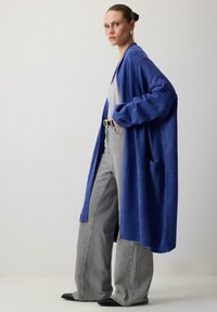 Naine, kes kannab üleväikest sinist kardiganit, valget sotti, laiad hallid teksad, musti jalanõusid ja kuldseid kõrvarõngaid, seisab puhtas valges seinas.