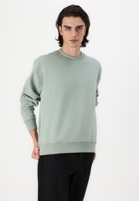 Hanorac verde deschis dintr-un material moale, cu guler rotund, manșete ribbate și o croială lejeră. Asortat cu pantaloni negri.