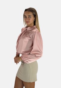 Zartrosa Satinbluse mit Puffärmeln und einer Schleife am Hals, kombiniert mit einem eng anliegenden, hellbeigen Minirock. Durchgehend glatte Textur.