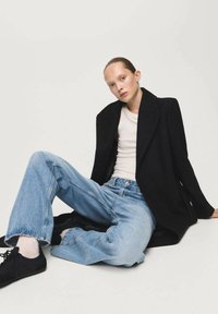 Zwarte wollen jas over een wit T-shirt, gecombineerd met lichtblauwe wijde jeans. Model zittend, draagt zwarte schoenen, tegen een minimalistische achtergrond.