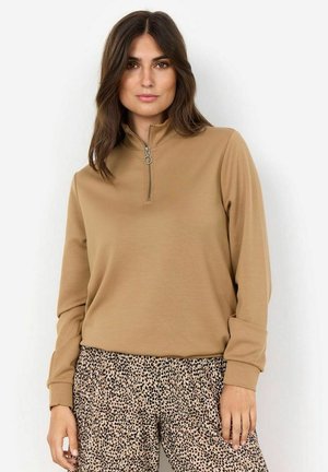 Beiger Zip-Pullover mit hohem Kragen und langen Ärmeln; kombiniert mit hellbraunen und schwarzen Leopardenmuster-Hosen. Glatte Stoffqualität.