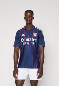 Tamsiai mėlynas futbolo marškinėliai su raudonomis vertikaliomis juostelėmis, puikuojasi baltu "Emirates" logotipu ir "OL" herbu. Trumpomis rankovėmis ir V formos kaklo dizainas.