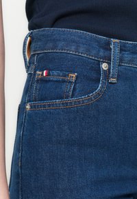 Dongeribukser med høy midje i mørkeblått, med gullsting, en liten frontlomme med røde, hvite og blå detaljer, samt en metallknapp-lukking.