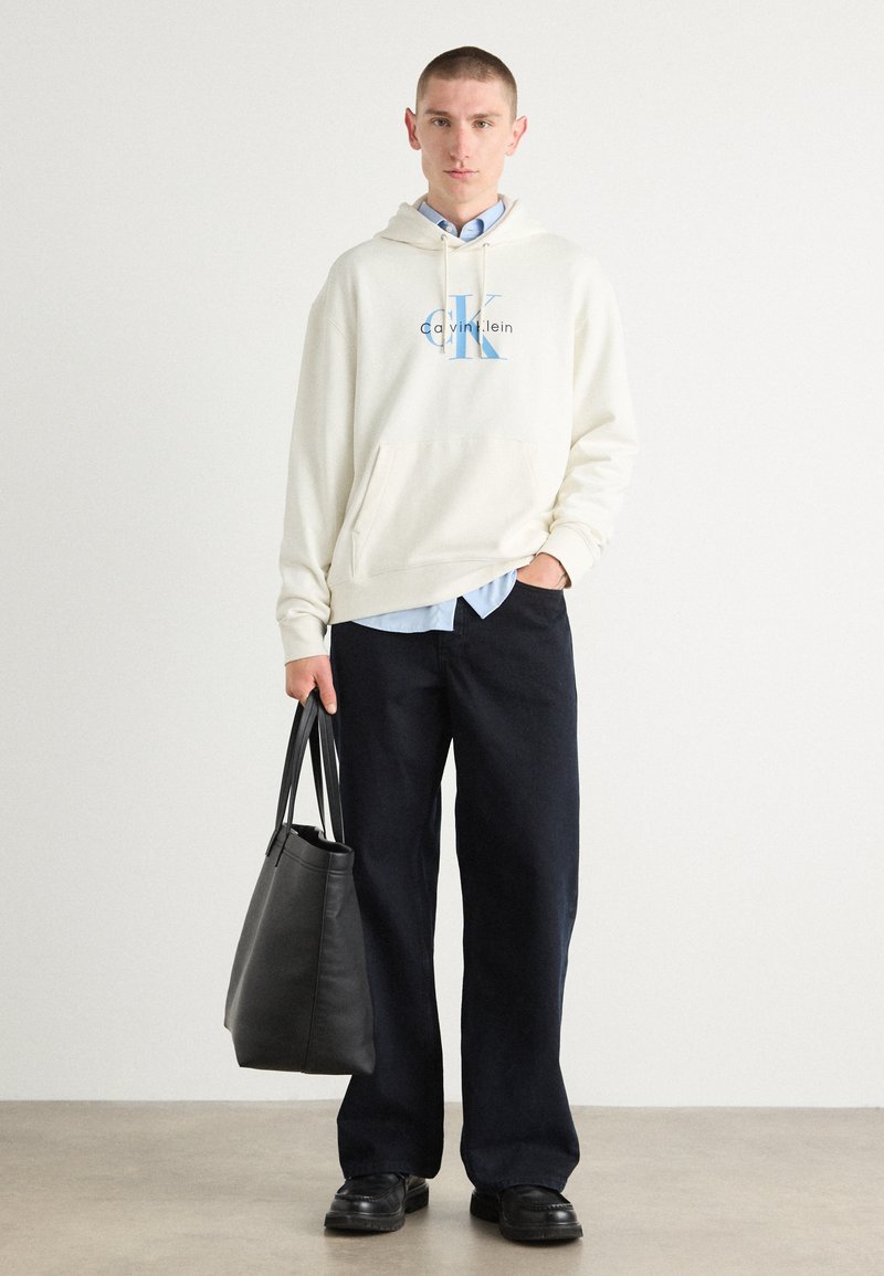 Felpa leggera color crema con logo blu, abbinata a una camicia azzurra chiara, pantaloni larghi neri e una borsa a tracolla in pelle nera.