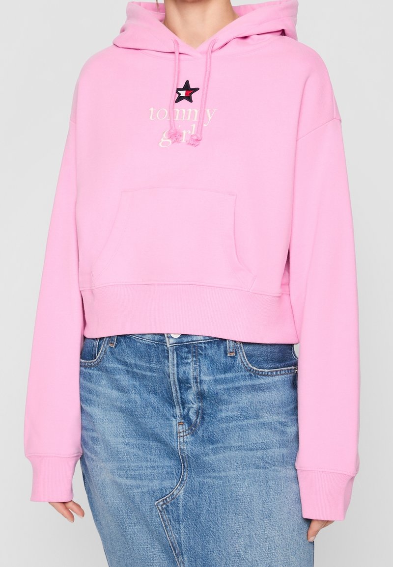 Personne portant un sweat à capuche court rose avec le texte "tommy girl" et un emblème étoile, associé à une jupe en jean bleue avec des coutures visibles.
