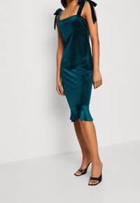 Teal sammetsklänning i midi-längd med fyrkantig halsringning, knytband och volangfald. Buren med svarta sandaler med öppen tå, vilket framhäver en slank silhuett.