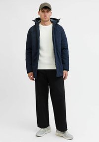 Giacca esterna blu navy con cappuccio, maglione bianco a coste e pantaloni larghi neri. Sneaker grigie completano il look. Design minimalista.