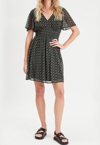 Robe à motifs floraux noirs avec manches courtes transparentes et encolure en V, dotée d'une taille smockée et d'une jupe évasée. Portée avec des sandales noires.