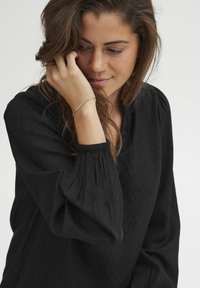 Juoda bluzonė su tekstūruotu audiniu, pasižyminti ilgomis rankovėmis su rauginėtomis rankogaliais. Modelis laiko ranką šalia veido, demonstruodama švelnų auksinį apyrankę.