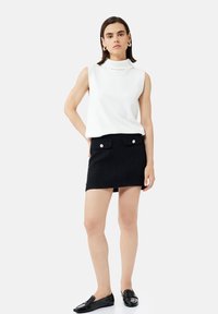 Top sin mangas de cuello alto blanco combinado con una mini falda negra texturizada que presenta dos bolsillos frontales y acentos de botones plateados. Mocasines negros.