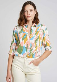 Chemise à manches courtes colorée avec des motifs de feuilles abstraites audacieux en rose, vert, bleu et jaune sur un fond clair ; tissu léger.