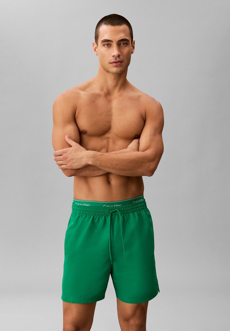 Homme musclé aux cheveux courts, bras croisés, portant un short de bain vert avec la ceinture Calvin Klein visible, sur fond gris.