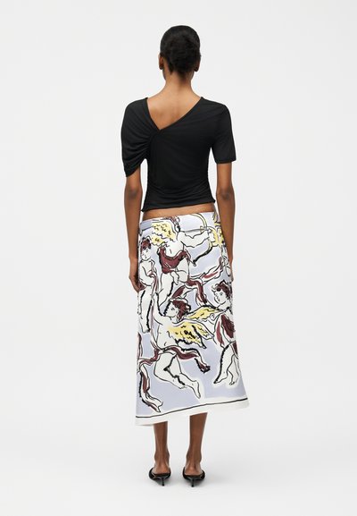 Γυναίκα με μαύρο ασύμμετρο τοπ και λευκή μίντι φούστα με κίτρινα και κόκκινα τυπώματα ερωτόκριτου, φωτογραφημένη από πίσω σε λευκό φόντο.