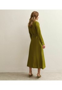 Abito verde oliva a maniche lunghe con vita aderente e lunghezza midi. Realizzato in tessuto testurizzato, presenta una chiusura a bottoni nascosta e polsini.