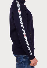Mörkblå zip-up sweatshirt med hög krage, har en vit sidosträng med "U.S. POLO ASSN." och tryck av den amerikanska flaggan. Texturerad tyg, avslappnad passform.