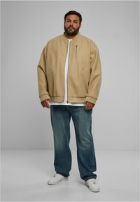 Jachetă bomber bej cu fermoar frontal, două buzunare laterale, croială lejeră, asociată cu blugi denim albaștri deschiși și adidași albi.
