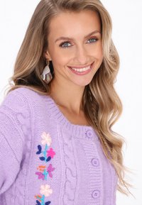 Levendula színű, kötött kardigán, fonott mintával, rózsaszín és kék virágos hímzéssel, valamint lila gombokkal az elején.