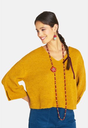 Frau mit dunkel geflochtenem Haar, die einen senfgelben Pullover, blaue Hose, rote Perlenkette und passende rote und blaue Ohrringe trägt, lächelnd nach links schauend.
