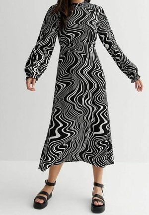 Kobieta w długiej sukience midi z czarno-białym falowanym wzorem, z długim rękawem, nosząca czarne sandały na platformie z paskiem wokół kostki, na jednolitym tle.