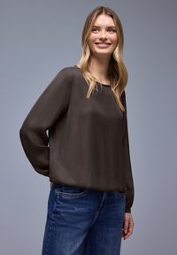 Blouse marron foncé à manches longues avec un col rond, fabriquée en tissu lisse, associée à un jean bleu, présentée sur un fond neutre.