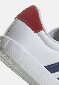Sneaker bianca con tomaia in sintetico testurizzato, accentuata da un tallone in suede rosso e una striscia blu navy. Suola in gomma con un motivo di adesione testurizzato.