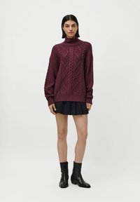 Kvinne iført en lilla ribbestrikket høyhalset genser, svart plisserte mini-skjørt og svarte skinnboots til midten av leggen som står foran en hvit bakgrunn.