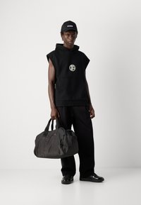 Young man wearing black sleeveless hoodie and cap, holding large black duffel bag, standing against a plain light background.