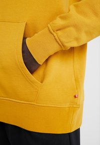 Main à peau foncée glissée dans la poche avant d’un sweat-shirt jaune moutarde avec poignet côtelé et petite étiquette rouge sur la couture latérale.