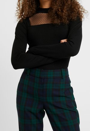 Personne aux cheveux bouclés portant un haut noir à manches longues avec un panneau transparent à pois et un pantalon à carreaux verts et bleu marine, les bras croisés.