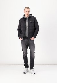 Chaqueta acolchada negra sobre un hoodie gris, combinada con jeans grises y zapatillas blancas. La chaqueta cuenta con un cierre de botones a presión y un bolsillo en el pecho.