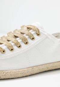 Bílé plátěné boty s zlatými pletenými tkaničkami a zlatými oky. Dokončeno pletenou jutovou podrážkou. Jednoduchý, čistý design.