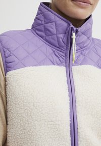 Vest din fleece cu partea superioară lavandă, cu un tip de matlasare în diamant, și partea de jos moale, crem. Are un guler înalt și un fermoar cu accente galbene.