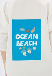 Biała koszula z niebieską grafiką i napisem "OCEAN BEACH" oraz kolorowymi ilustracjami meduz, kraba, muszli i rozgwiazdy.