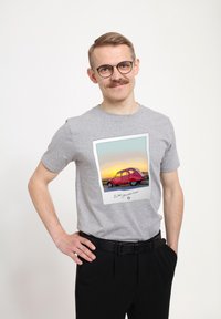 Šedé bavlněné tričko s potiskem červeného vintage auta na pozadí západu slunce, orámované jako fotografie.