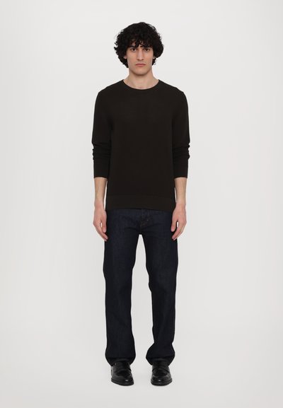 Dunkelbrauner, gestrickter Pullover mit langen Ärmeln, kombiniert mit dunkelblauen Jeans und schwarzen Schuhen, Model steht vor einem weißen Hintergrund.