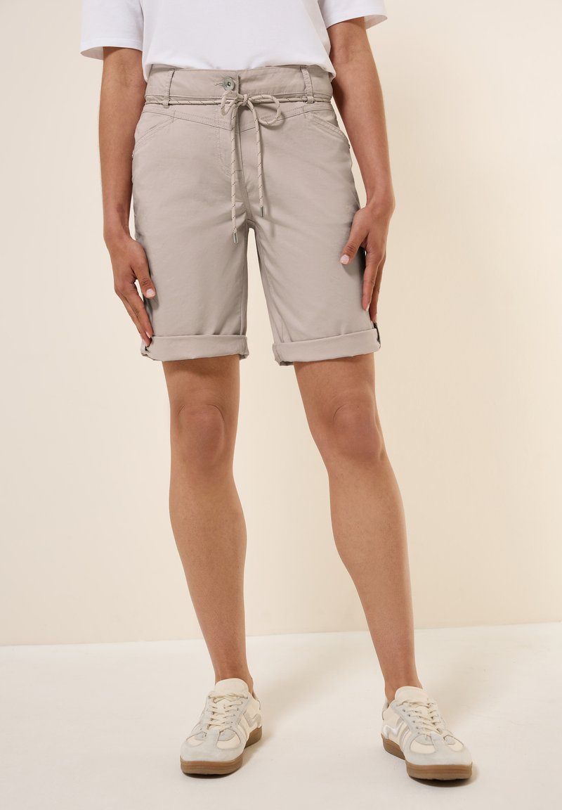 Pantaloni corti beige con vita regolabile, orli arrotolati e tasche laterali, realizzati in tessuto leggero. Abbinati a sneaker bianche.
