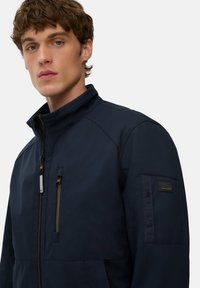 Ung mand med krøllet brunt hår iført en marineblå zip-up jakke med flere lynlåslommer og en høj krave.