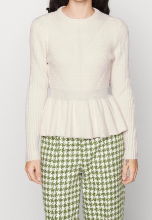 Femme portant un pull côtelé crème à basque et un pantalon à motif pied-de-poule vert et blanc, debout devant un fond uni.