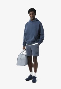 Felpa con cappuccio blu navy con tasca frontale, abbinata a pantaloni corti coordinati. Borsa grigia chiara con zip e manico. Il modello indossa scarpe blu navy e calze bianche.